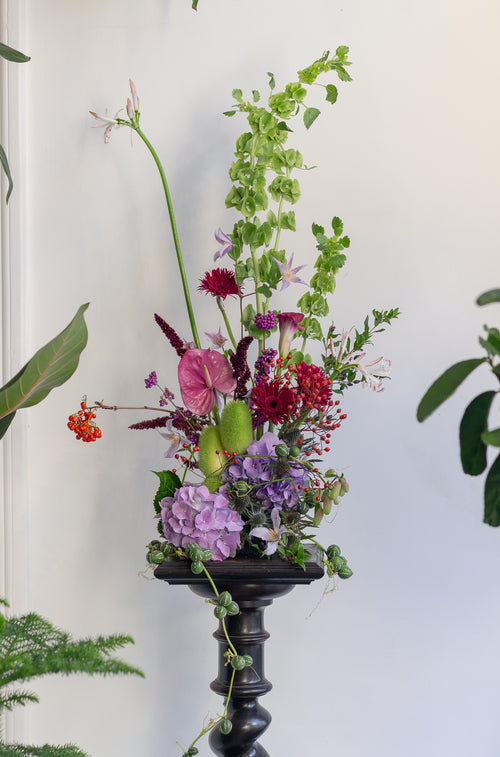 A lush floral arrangement featuring hydrangeas, anthuriums, roses, and green foliage in a tall black pedestal vase, set against a clean white wall.