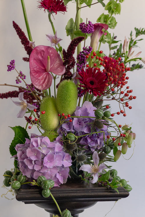 A vibrant and lush floral arrangement featuring pink and purple hydrangeas, red gerbera daisies, green seed pods, and red berries, presented in a dark wooden pedestal bowl against a plain light background.